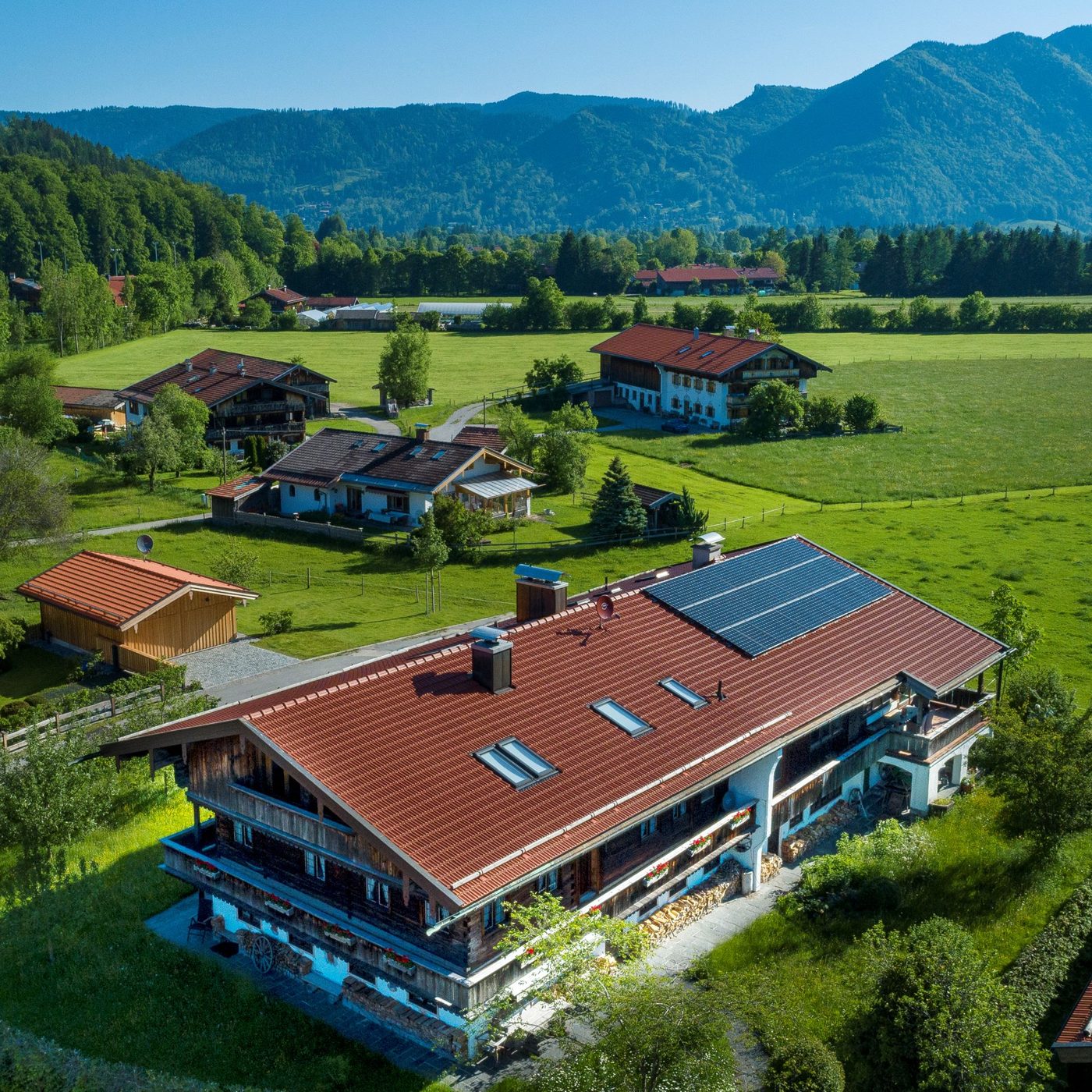 Luftbild mit dem Hof im Vordergrund, dahinter Wiesen, Höfe und Berge